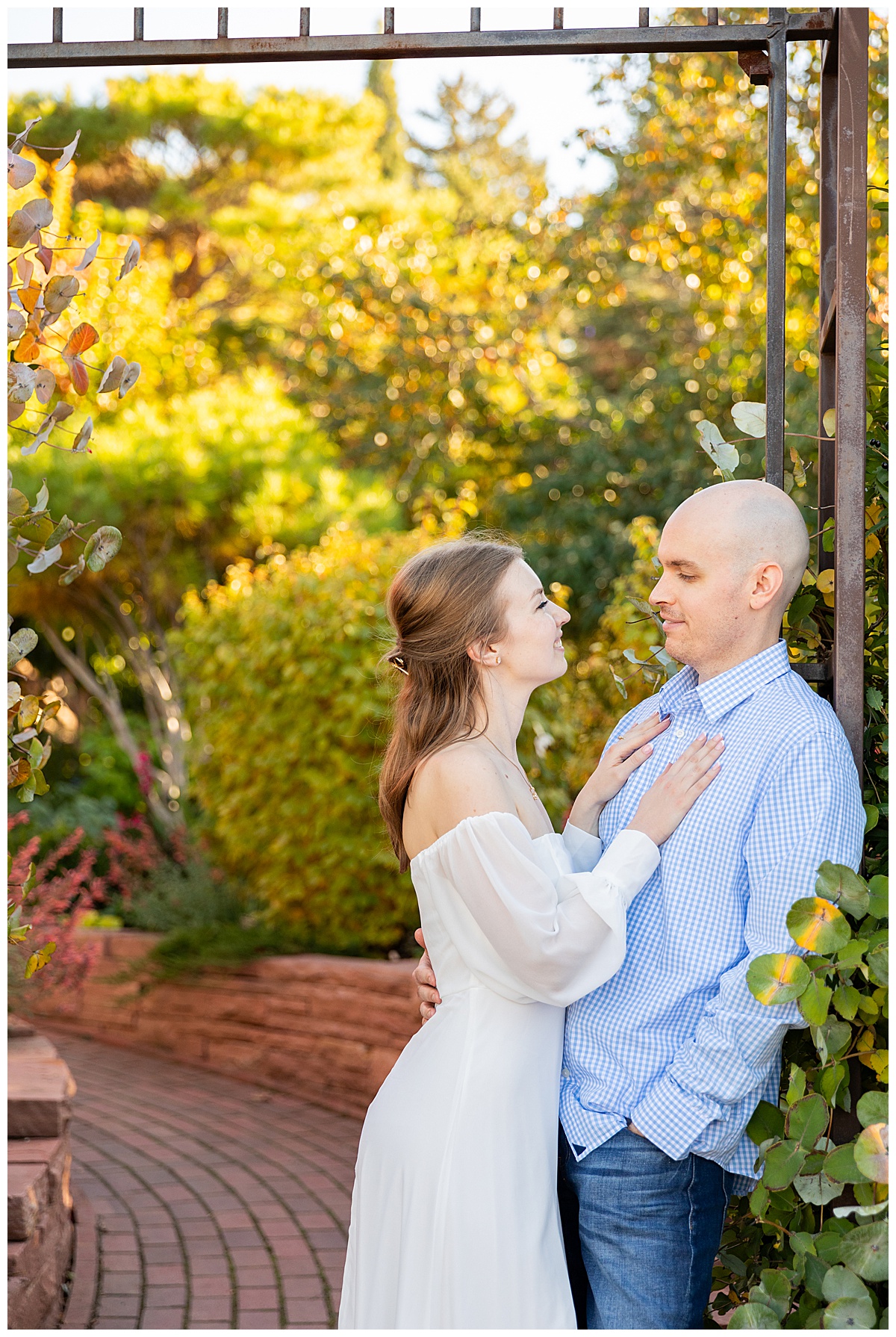 A couple poses for their botanic gardens engagement session. The blonde woman is wearing a white long sleeve dress and the bald man is wearing a blue plaid shirt and jeans.