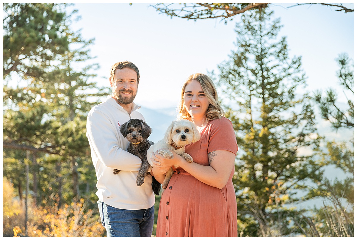 A couple poses for their Lookout Mountain couples session. The woman is wearing a terracotta linen dress and the man is wearing a white jacket and blue jeans. They are holding small fluffy dogs, one is black and one is white.
