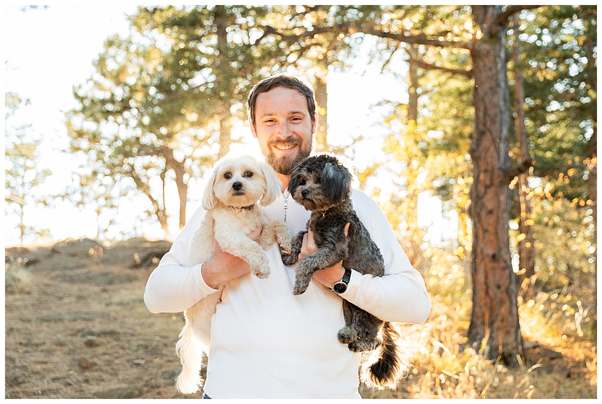 A man poses for photos. He is wearing a white jacket and blue jeans. He is holding both dogs.