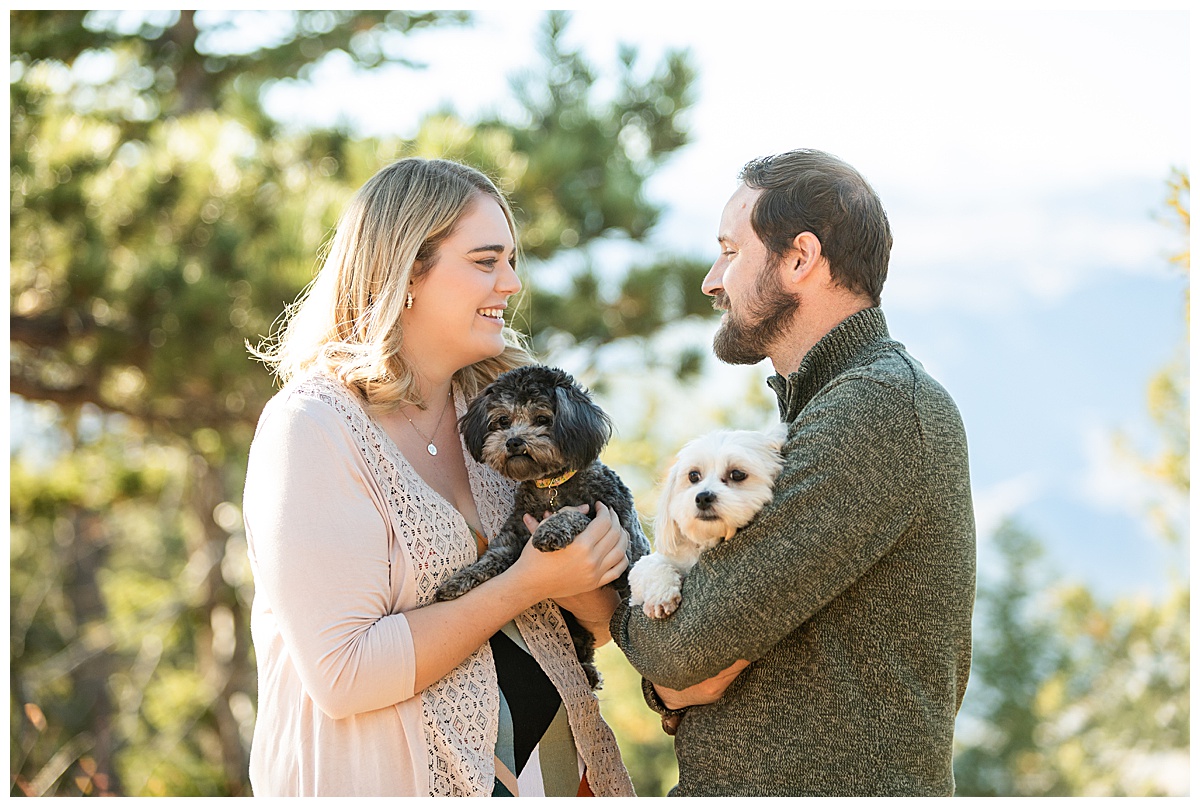 A couple poses for their Lookout Mountain couples session. The woman is wearing a geometric print shirt with a sweater and the man is wearing a green sweater and khaki pants. They are holding small fluffy dogs, one is black and one is white.
