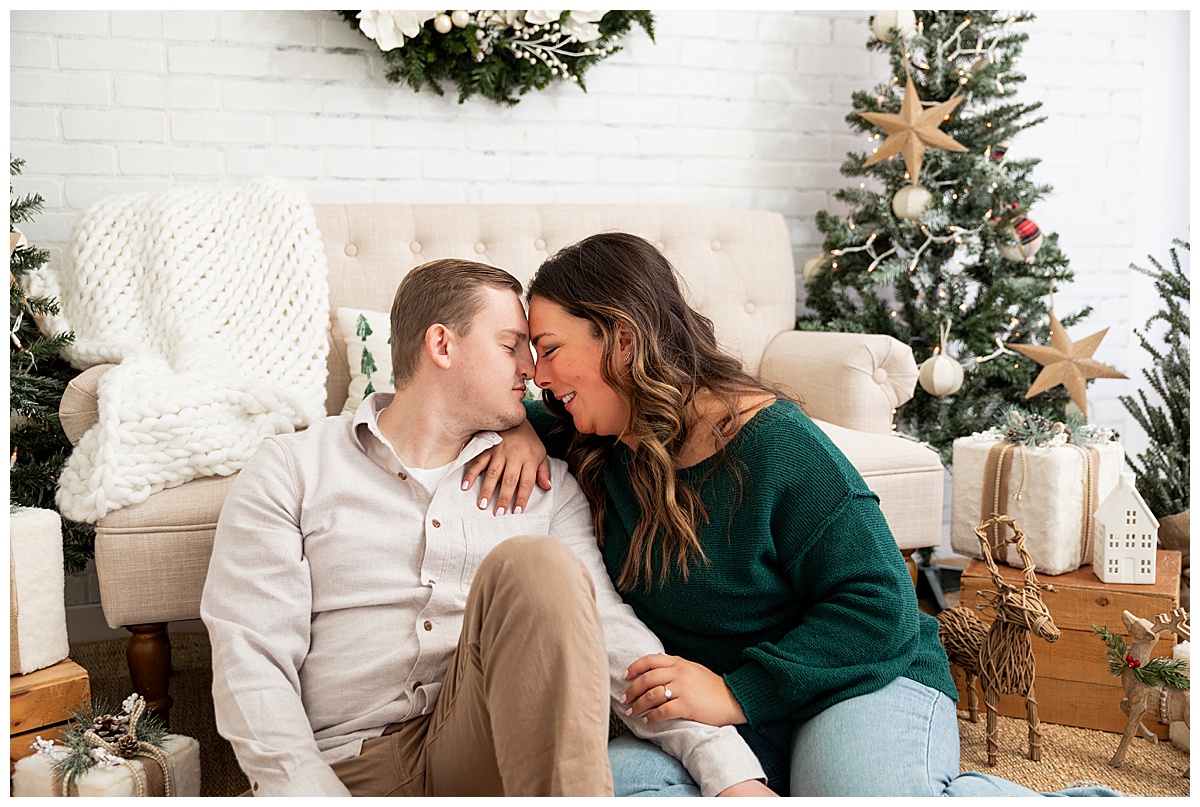 A couple poses in front of a tree, wreath, and couch. The woman is wearing a green sweater and jeans. The man is wearing a cream button down and khakis.