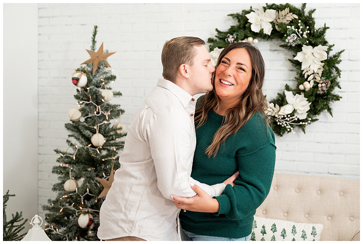 A couple poses in front of a tree, wreath, and couch. The woman is wearing a green sweater and jeans. The man is wearing a cream button down and khakis.