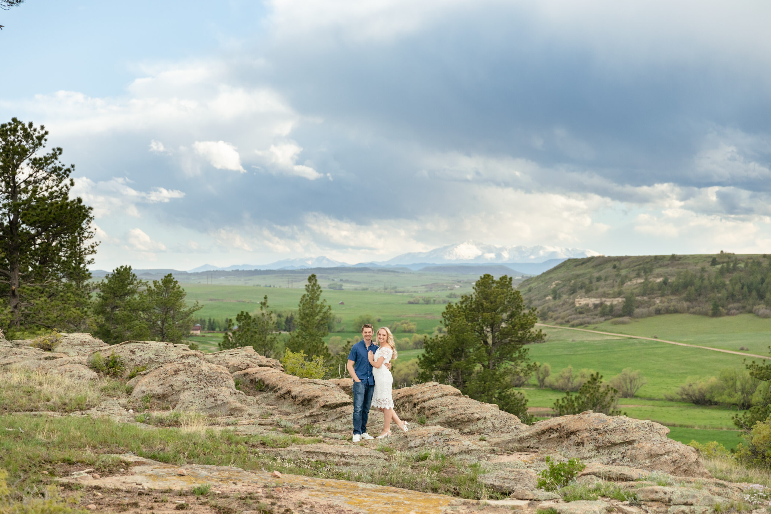 A couple poses on a rock cliff in front of green fields and mountains in the far background. The man wears a blue short sleeve button down and jeans. The woman wears a mid-length lace white dress.