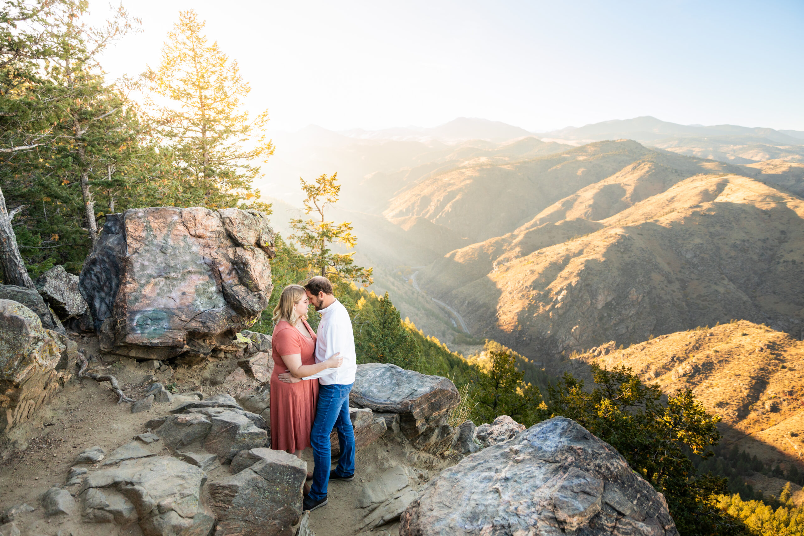 A man and woman stand on the edge of a cliff overlooking a mountain view. The woman is wearing a pink dress and the man is wearing a white sweater with blue jeans.