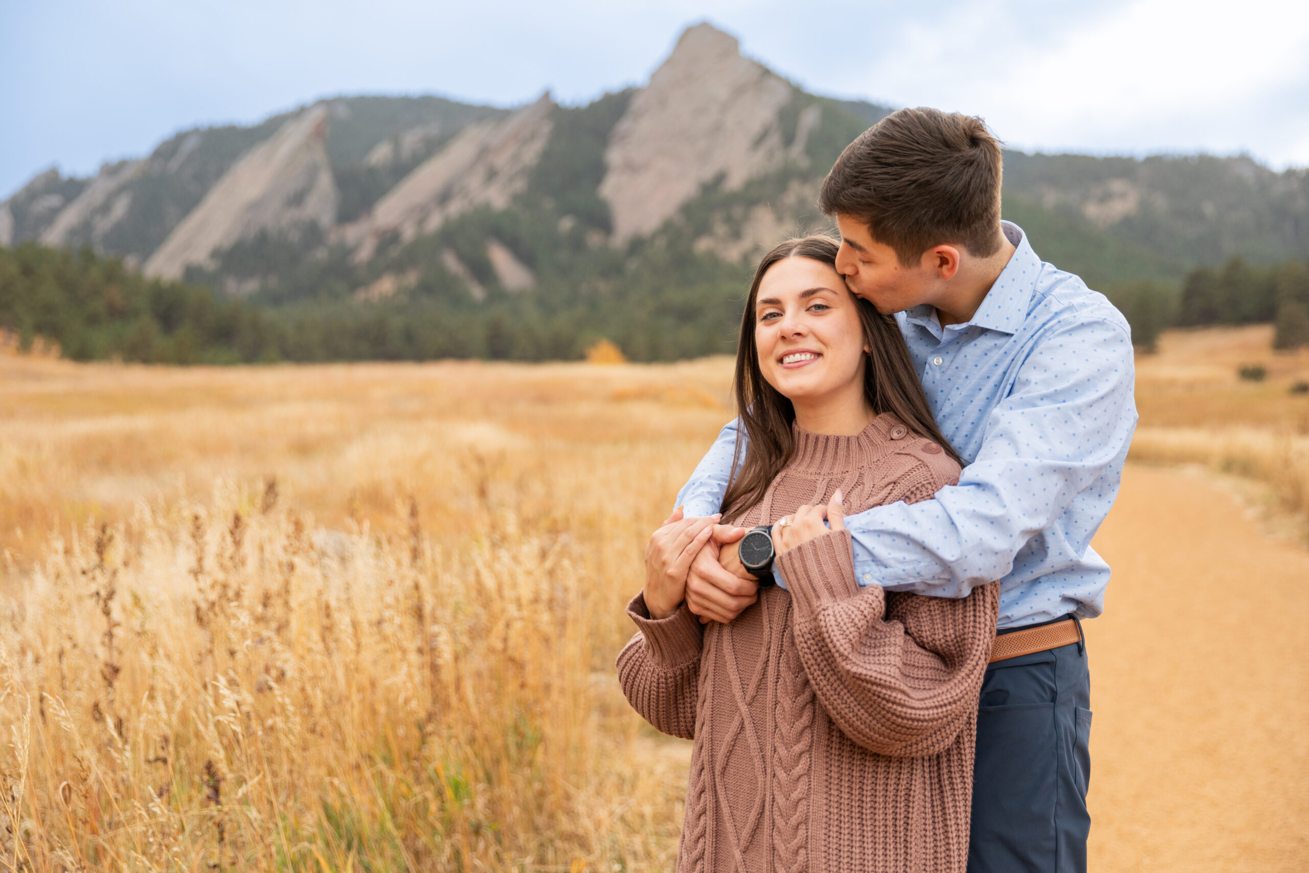 A couple cuddles on a path in front of a golden field and mountains in the background during their engagement session. The man is wearing a blue polka dot button down and blue pants. The woman is wearing a mauve sweater dress.