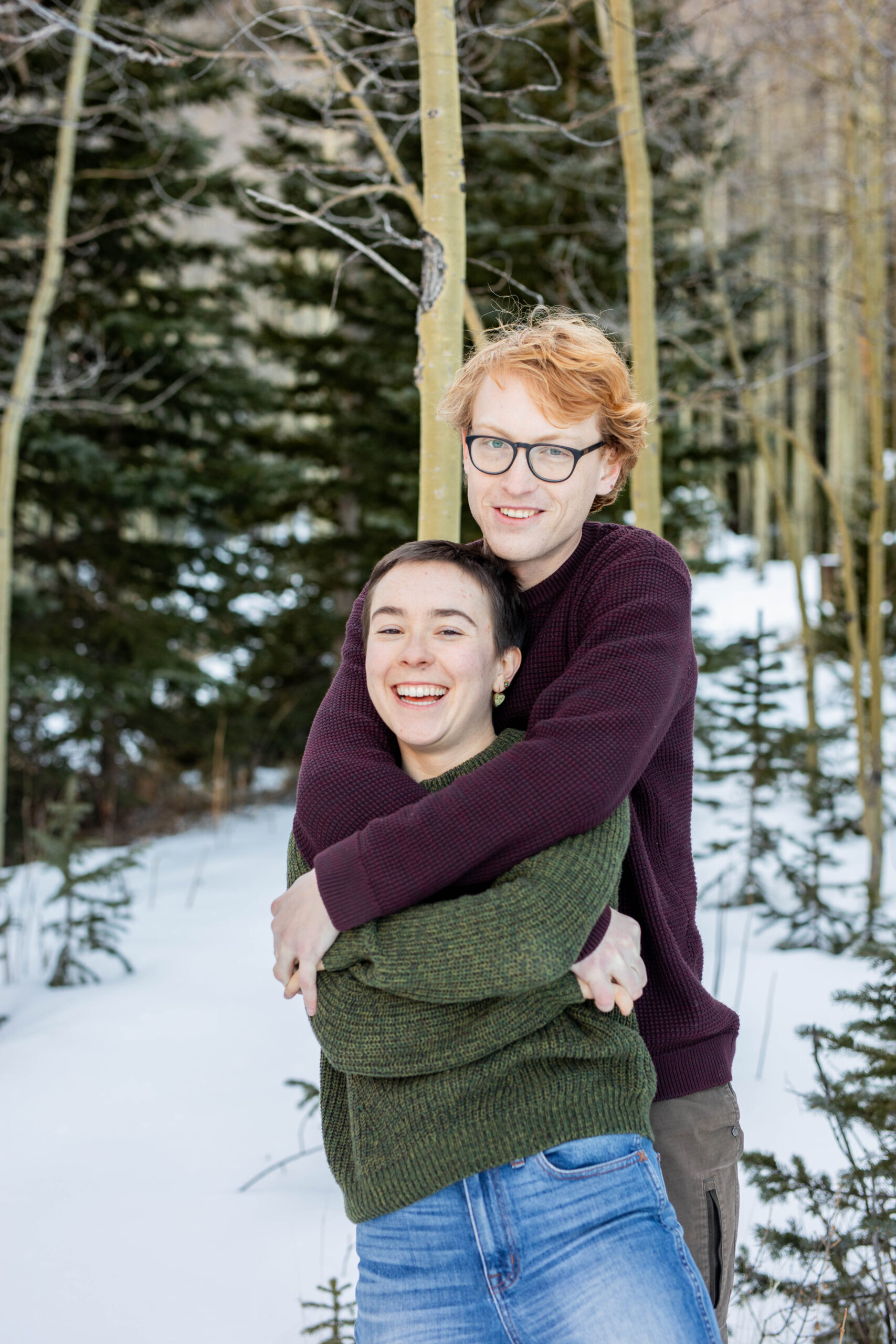 A couple cuddles in a forest of pine trees and aspens. The woman is wearing a green sweater and the man is wearing a red sweater.