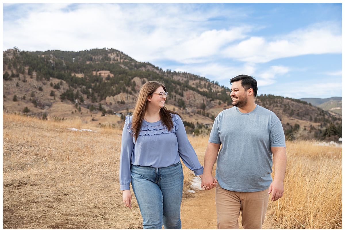 A couple poses in front of the Flatirons for their Boulder engagement session.  They are both wearing blue; the man has dark brown hair and a beard, the woman has long brown hair and glasses.