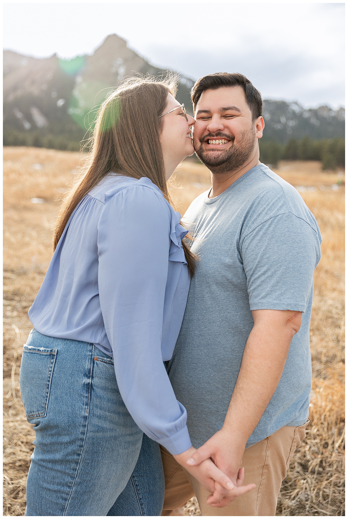 A couple poses in front of the Flatirons for their Boulder engagement session. They are both wearing blue; the man has dark brown hair and a beard, the woman has long brown hair and glasses.