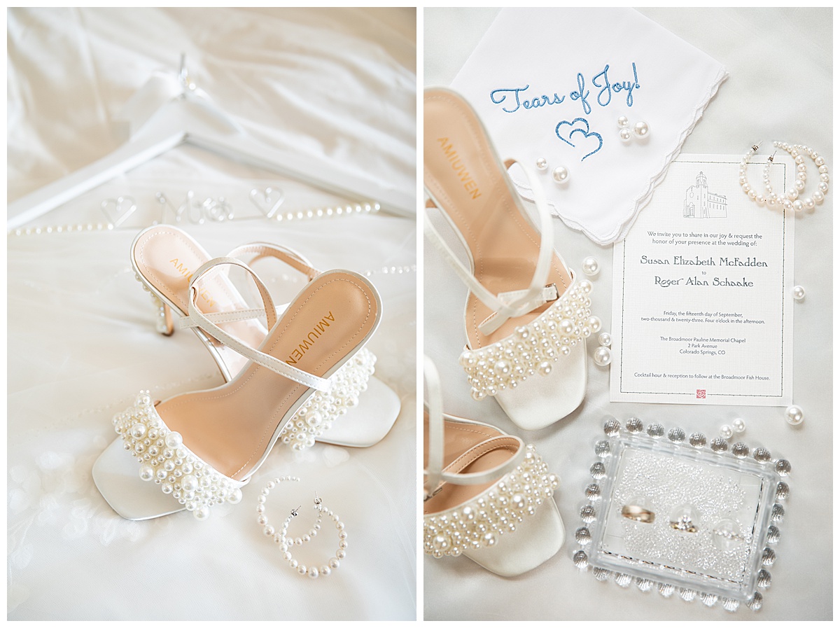 White wedding details: strappy heels with pearls, pearl hoop earrings, a "Mrs" hanger, a handkerchief with the words "Tears of Joy!" embroidered on it, a glass bead box with their rings, and a wedding invitation in black and white with the Broadmoor Resort outline on it.