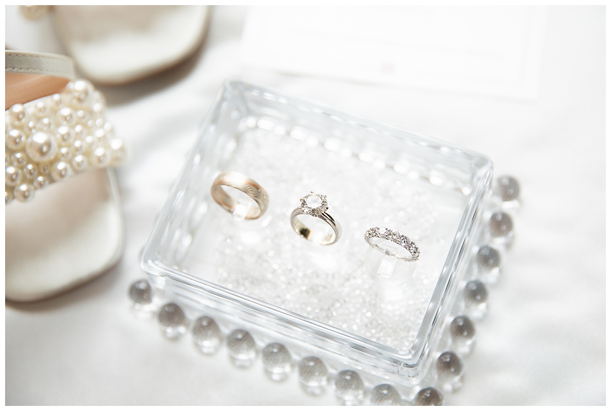 White wedding details: strappy heels with pearls, pearl hoop earrings, a "Mrs" hanger, a handkerchief with the words "Tears of Joy!" embroidered on it, a glass bead box with their rings, and a wedding invitation in black and white with the Broadmoor Resort outline on it.