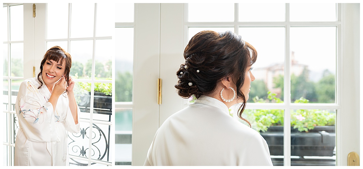 A bride with brown hair is putting her pearl hoop earrings in; she is wearing a white floral robe and her hair is in an updo