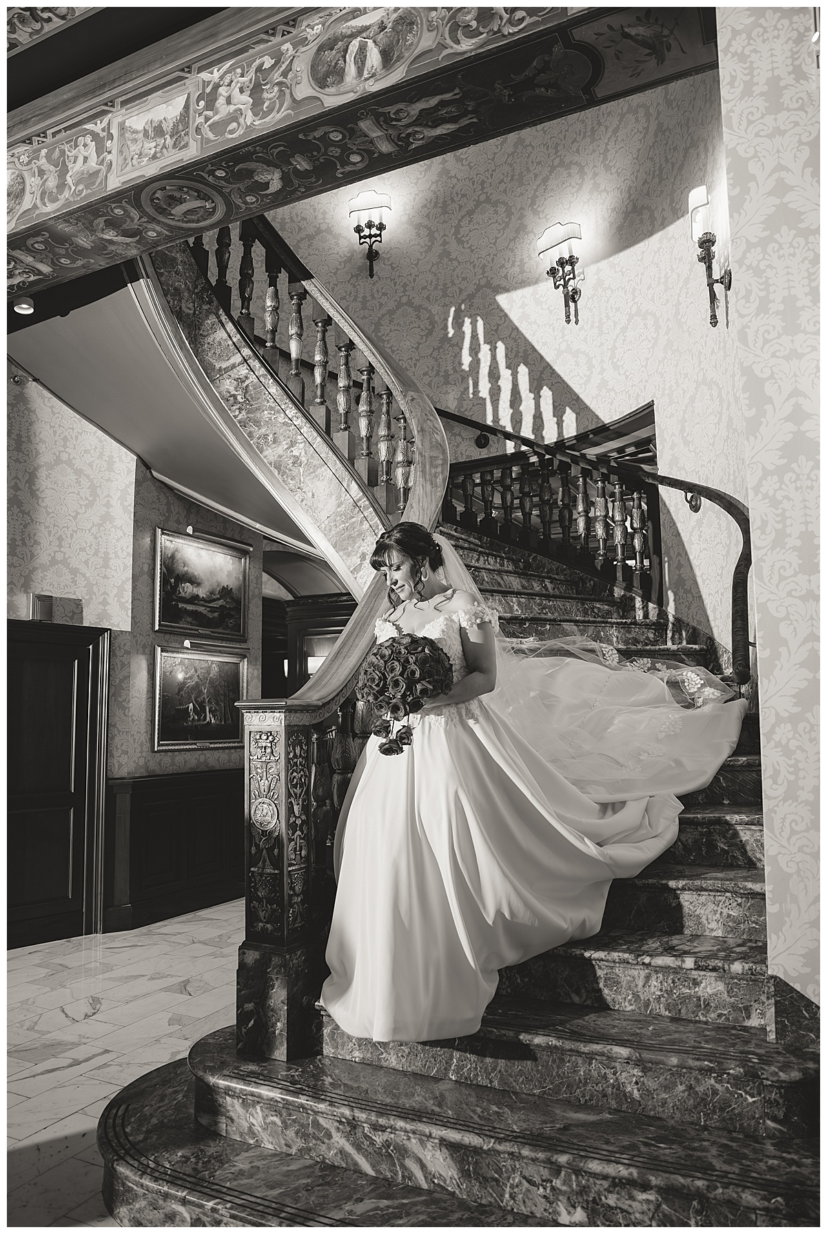 A black & white photo of the bride walking down the grand staircase. She is carrying a bouquet of red roses