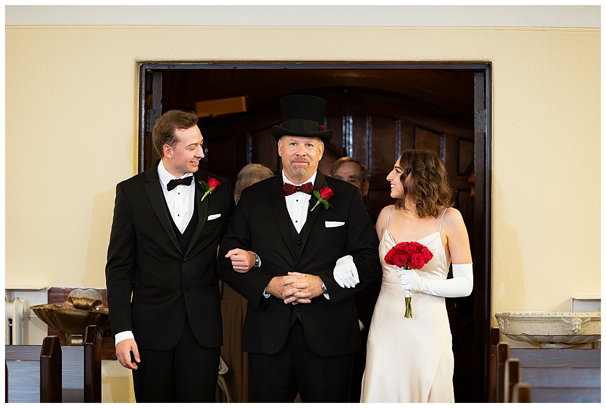 The groom walks down the aisle to the alter inside the wedding chapel for their ceremony. The groom is wearing a black suit. He is escorted by his son in a black suit and his daughter in a champagne silk dress.