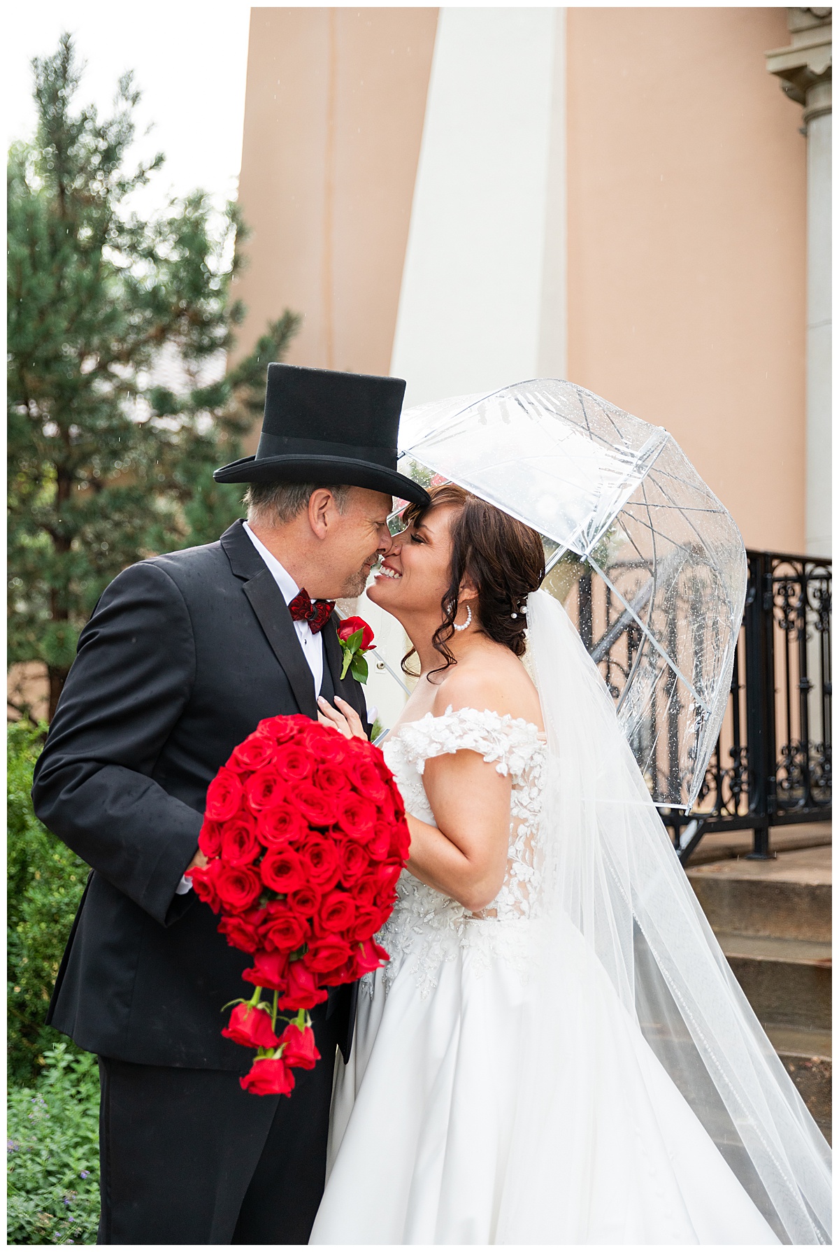 Couples portraits of a bride and groom during their Broadmoor wedding. The groom is wearing a black suit and black top hat. The bride is wearing a lace ballgown and a long veil. They are posing with a clear umbrella.