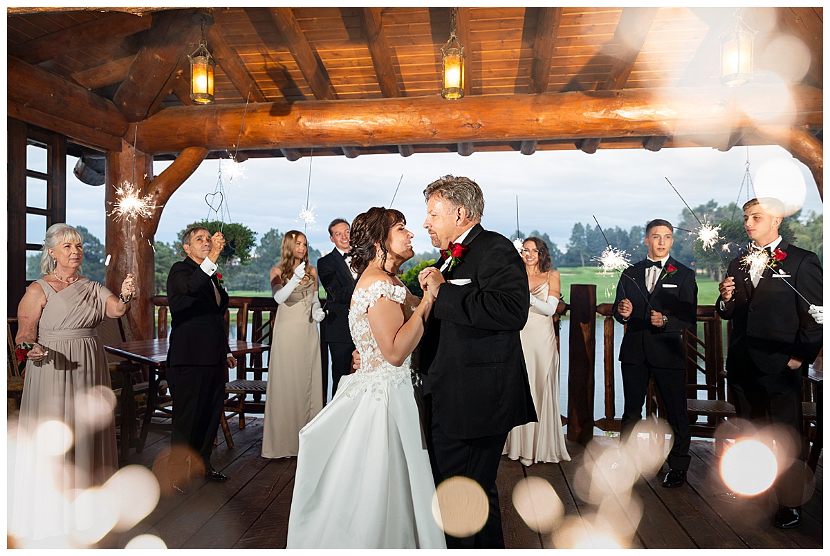 The bride and groom do their first dance on the back porch of the lake house. Their family are holding sparklers around them while they dance.