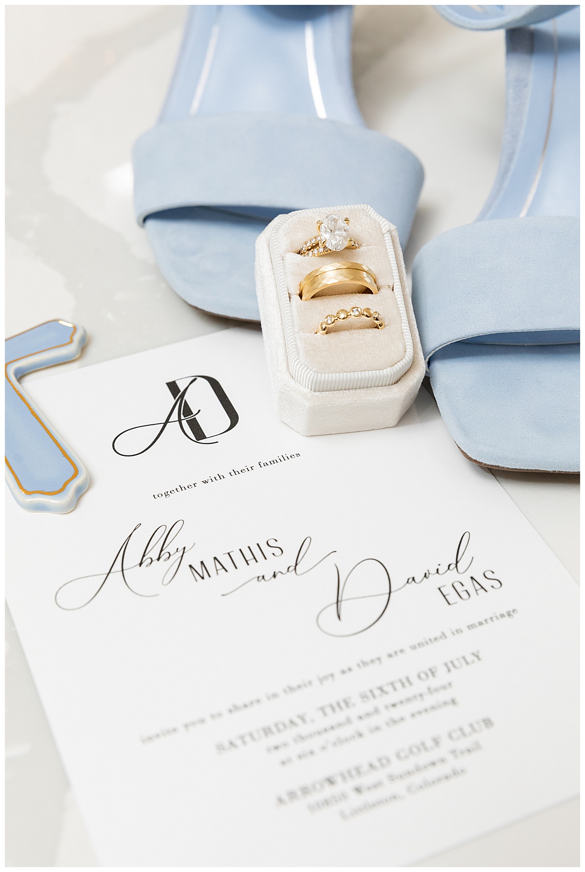 Top down images of a bride's wedding day details. There is a black and white wedding invitation, gold rings, gold and pearl earrings, blue suede shoes, and a light blue and gold ceramic cross.