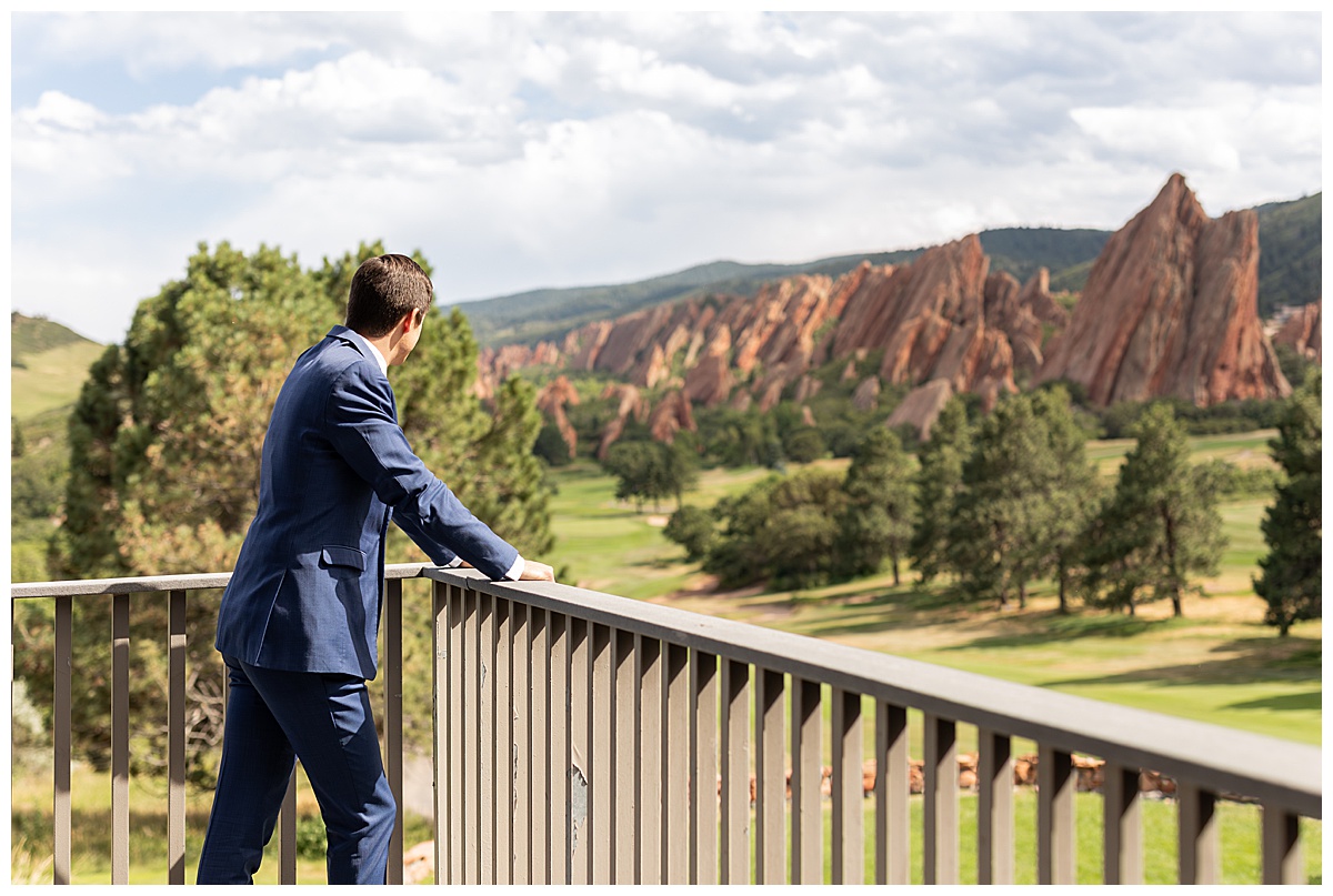 The groom is standing on a balcony leaning on the railing. He is looking over the golf course and red rocks in the background. He is wearing a navy blue suit.