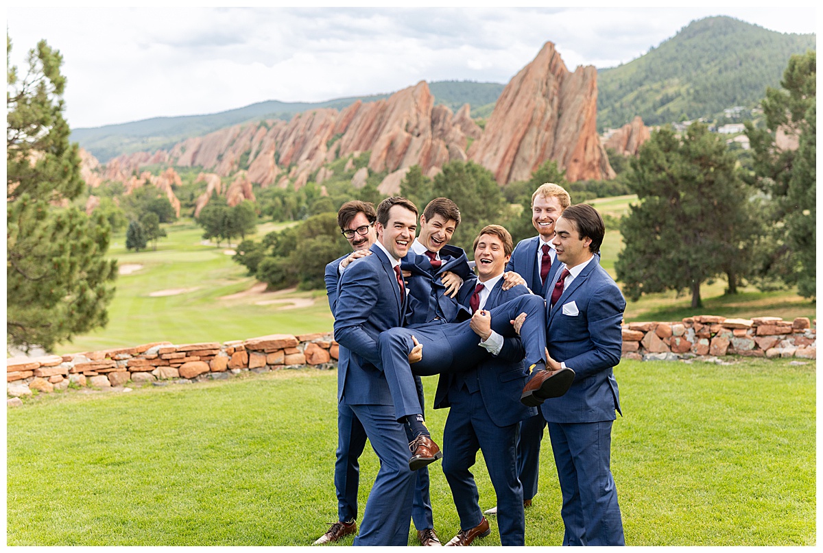 The groom is getting ready with his groomsmen. They are all wearing navy blue suits and maroon red ties. They are lifting him up.