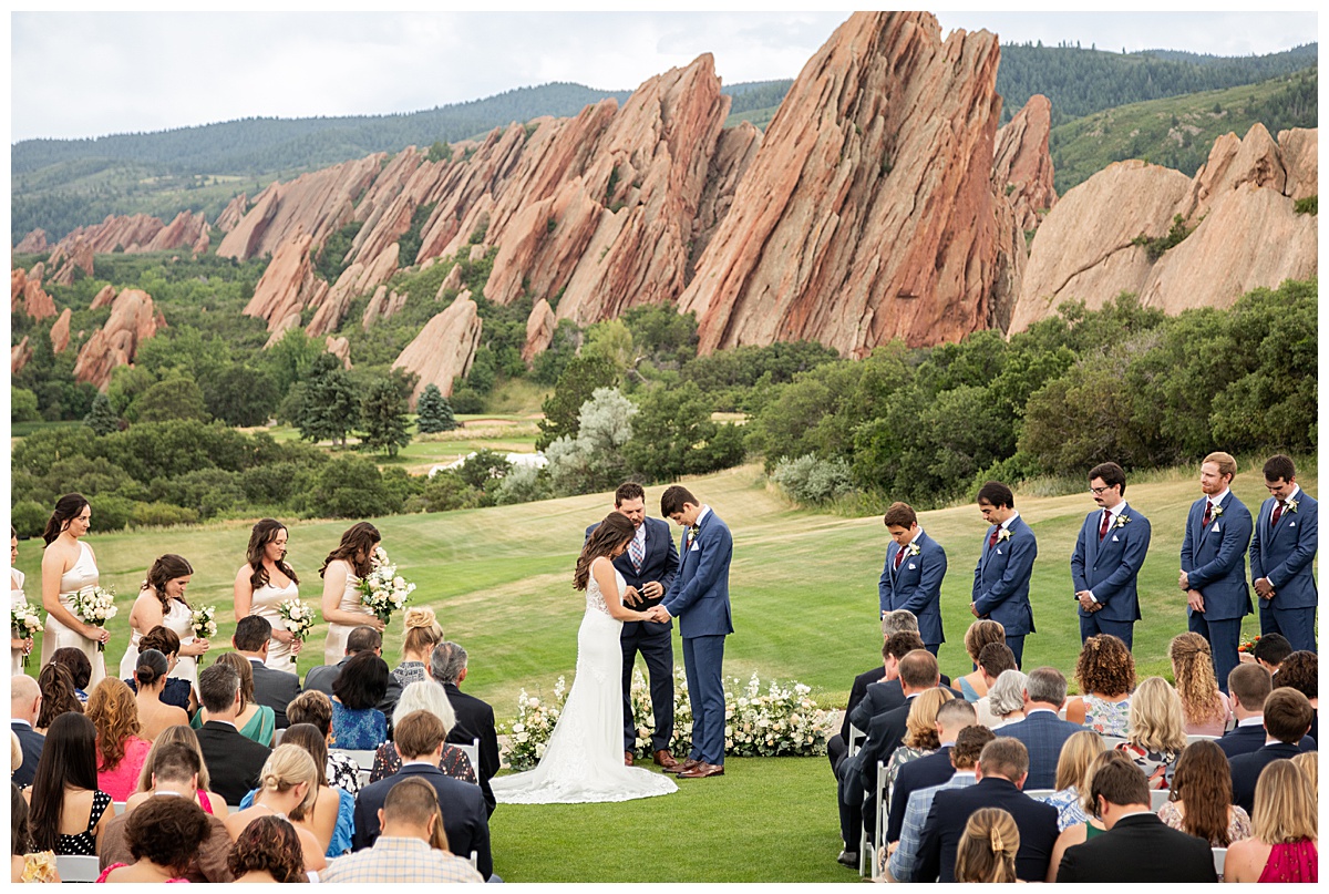 The couple hold hands during their wedding ceremony. Their bridal party is standing next to them and their guests are seated. The red rocks and golf course are in the background.