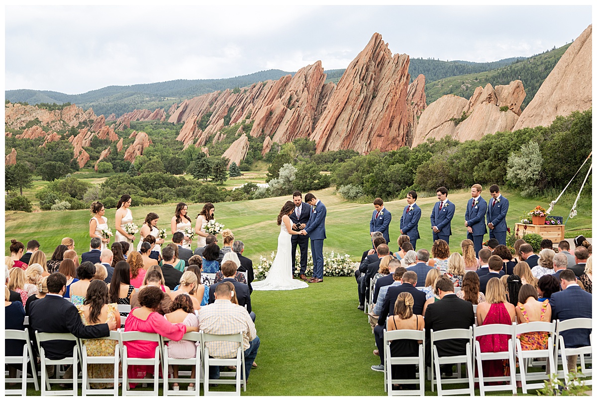 The couple hold hands during their wedding ceremony. Their bridal party is standing next to them and their guests are seated. The red rocks and golf course are in the background.