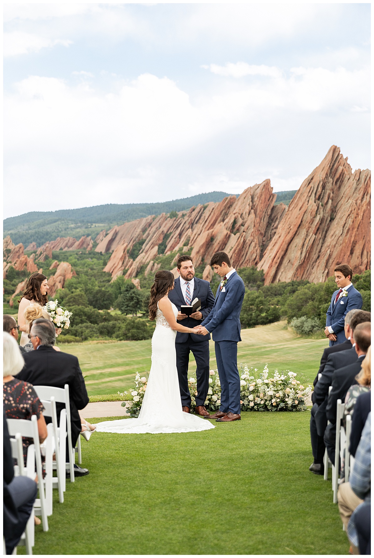 The couple hold hands during their wedding ceremony. Their bridal party is standing next to them and their guests are seated. The red rocks and golf course are in the background.