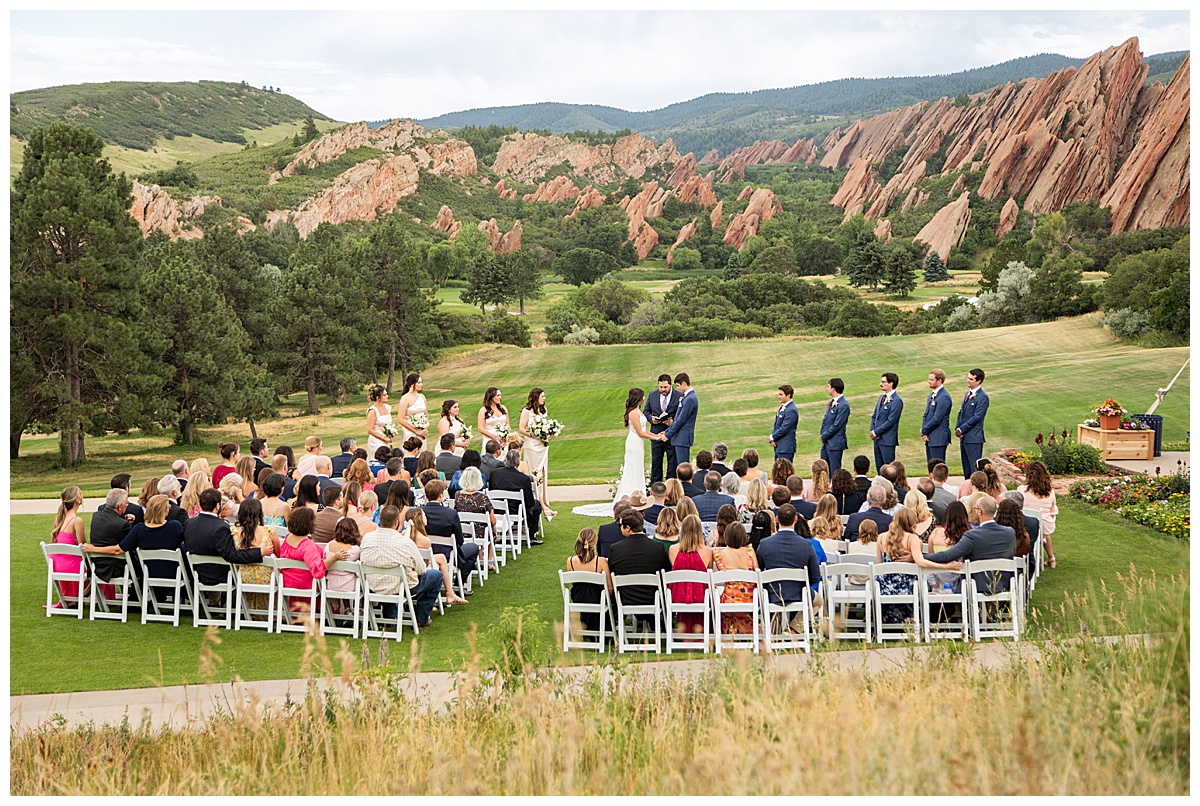 The couple hold hands during their wedding ceremony. Their bridal party is standing next to them and their guests are seated. The red rocks and golf course are in the background.