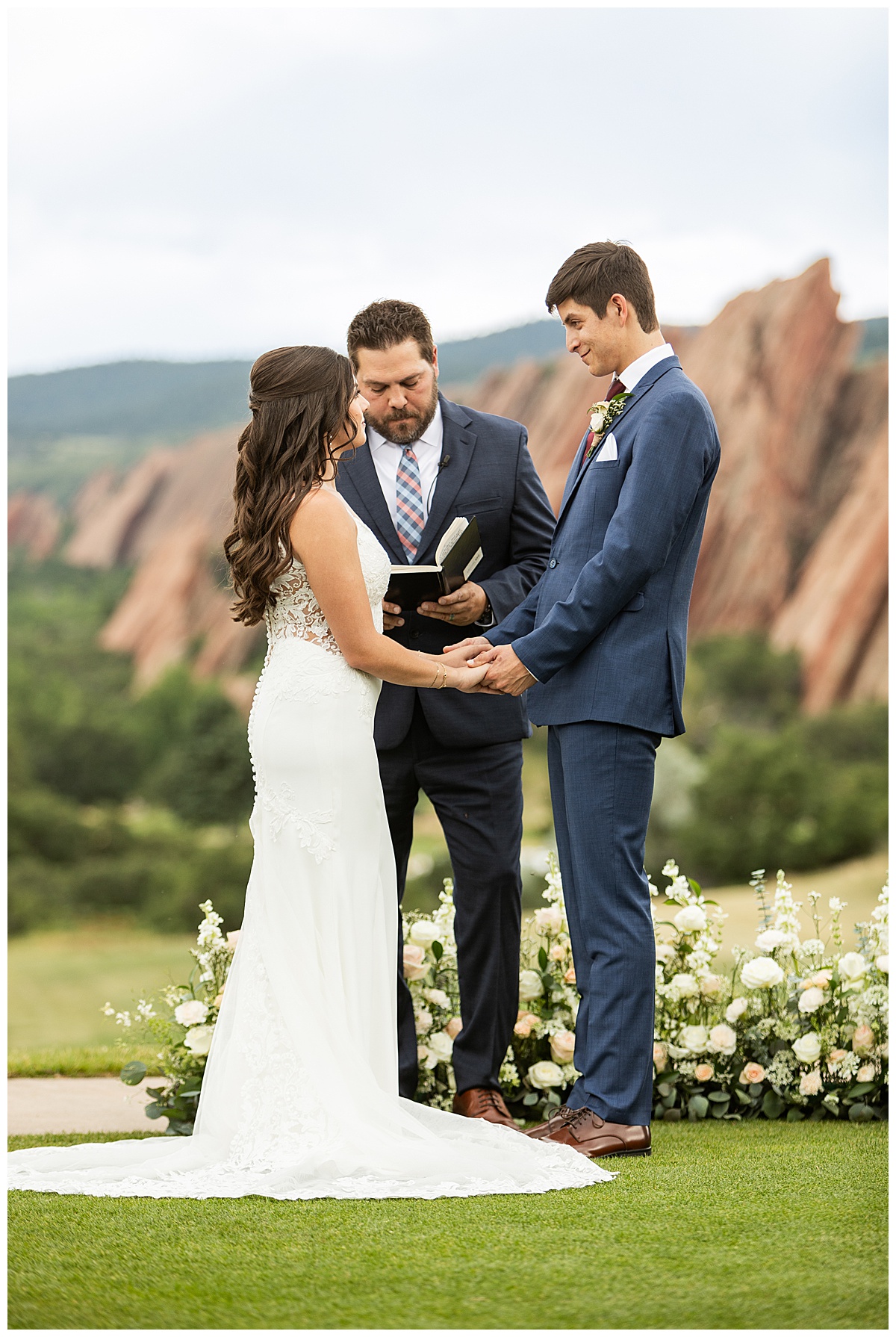 The couple hold hands during their wedding ceremony. Their bridal party is standing next to them and their guests are seated. The red rocks and golf course are in the background.