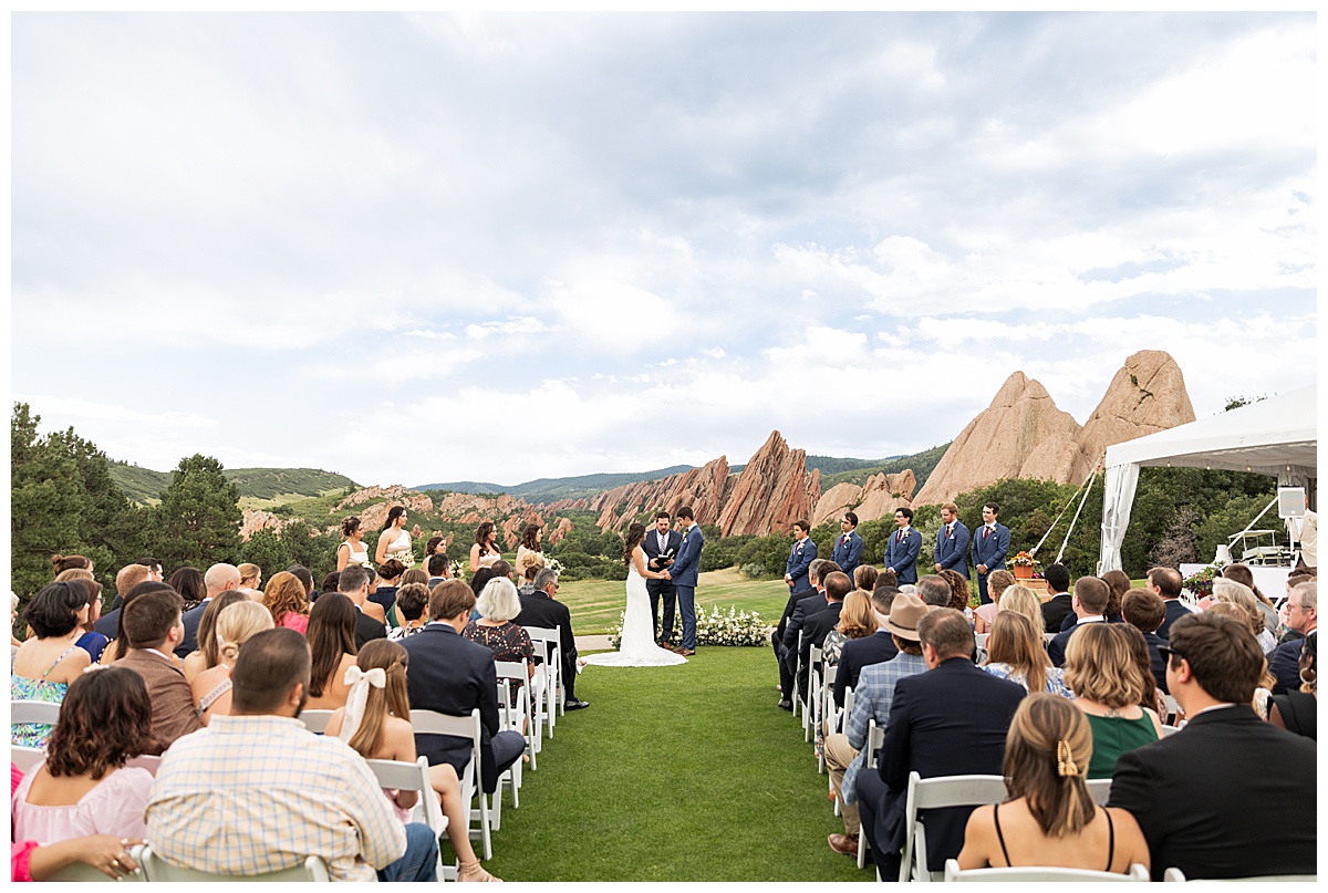 The couple hold hands during their wedding ceremony. Their bridal party is standing next to them and their guests are seated. The red rocks and golf course are in the background.