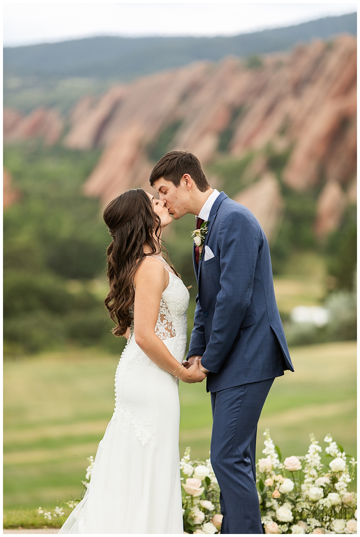 The couple have their first kiss during their wedding ceremony. Their bridal party is standing next to them and their guests are seated. The red rocks and golf course are in the background.