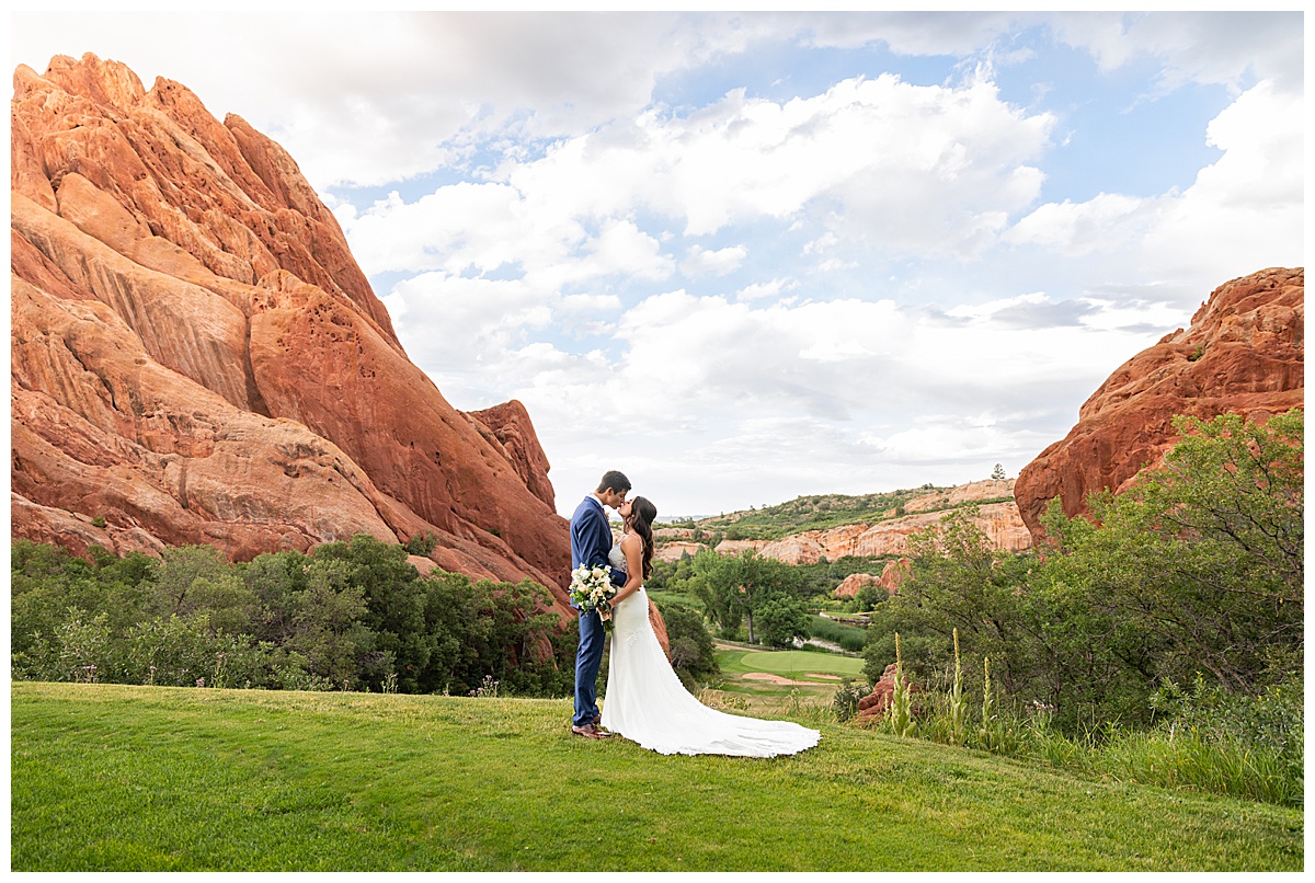 The couple pose on the golf course for bridal portraits. She is holding her bouquet. There are big red rocks in the background.