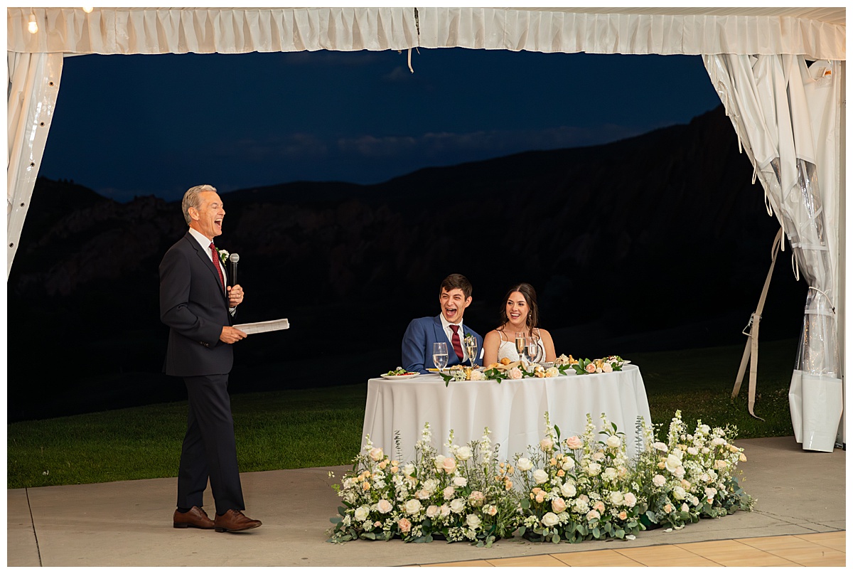 The bride and groom laugh during the father of the bride's toast. It is nighttime and they are under the reception tent.