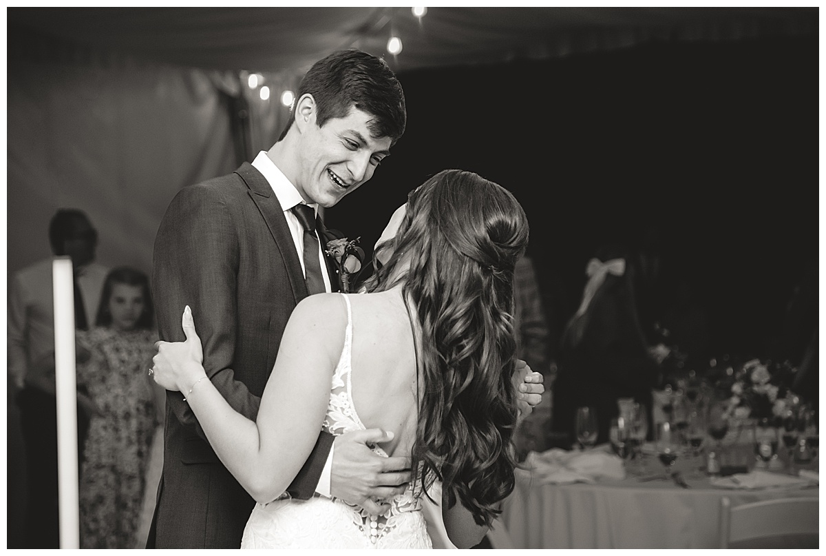 The bride and groom have their first dance. It is nighttime and they are under the reception tent. The photo is in black and white.