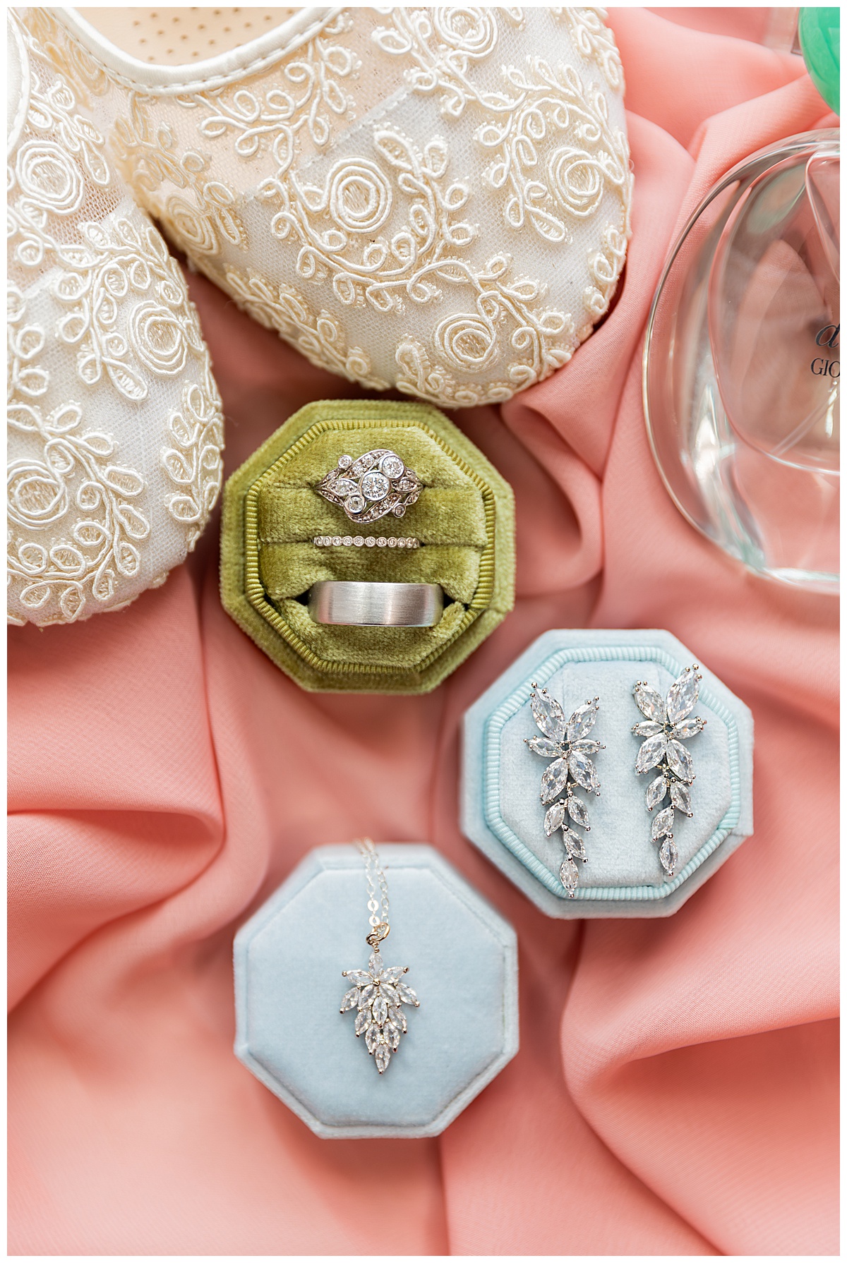 A close up photo of the couple's rings, the bride's jewelry, and her shoes. The rings and jewelry are in green and blue ring boxes. Everything is sitting on pink fabric