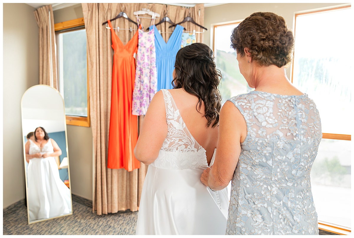 The bride's mom helps zip up her dress in front of a window and mirror. The bride's dress is white with lace and an a-line skirt