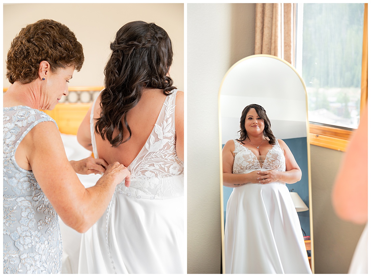 The bride's mom helps zip up her dress in front of a window and mirror. The bride's dress is white with lace and an a-line skirt