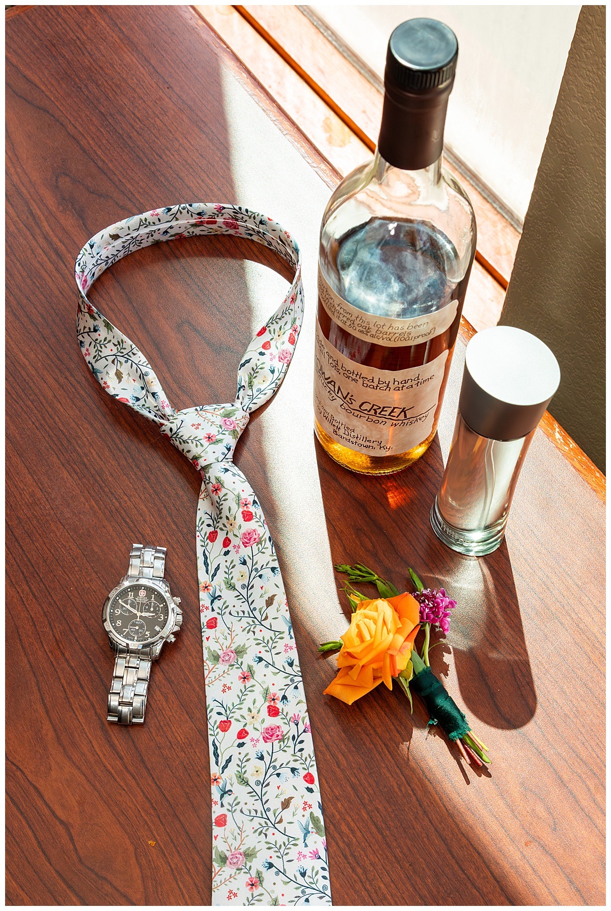 A close up photo of the groom's details. A floral tie, silver link watch, water bottle, orange rose boutonnière, and bottle of Rowan's Creek whiskey sit on a wood table. 