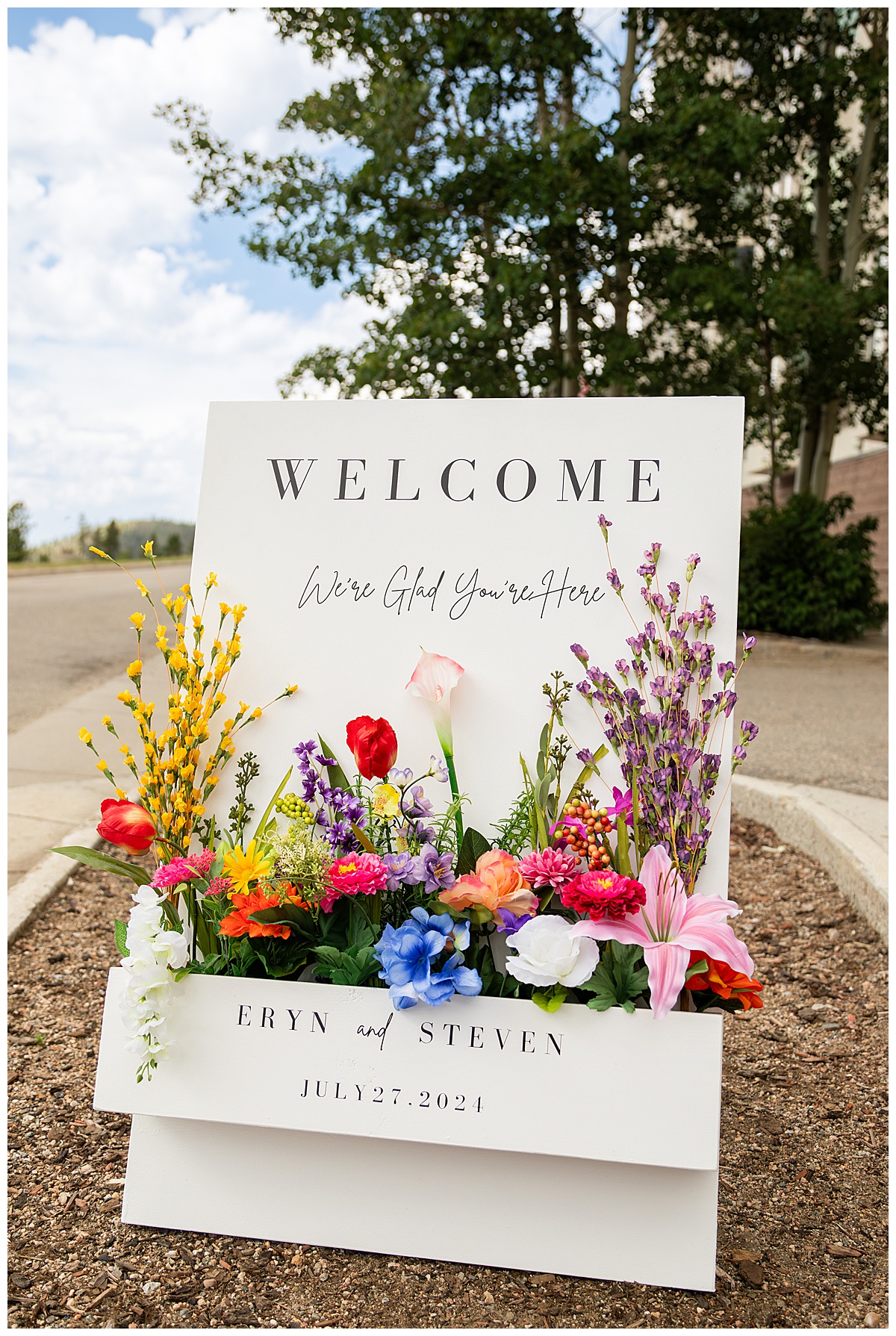 A close up photo of the couple's welcome sign. It is white with lots wildflowers. The sign reads "Welcome, we're glad you're here. Eryn and Steven. July 27, 2024"