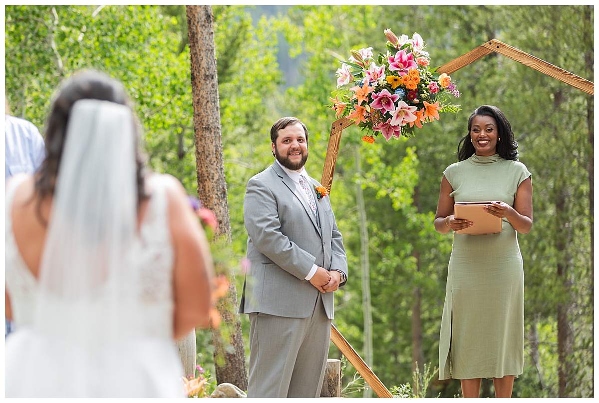 Photos of the groom reacting to the bride walking down the aisle. There is a big heptagonal arch and pine trees behind him.