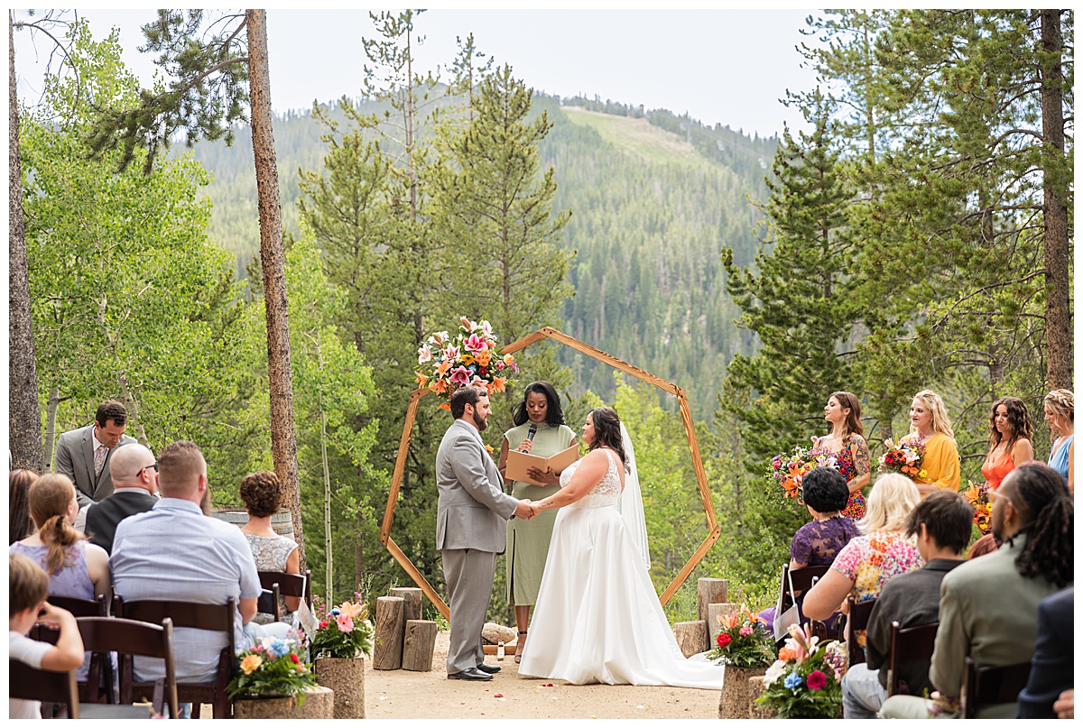 The bride and groom get married at Winter Park Mountain Lodge. They are holding hands. A heptagonal arch, pine trees, and a mountain are behind them.