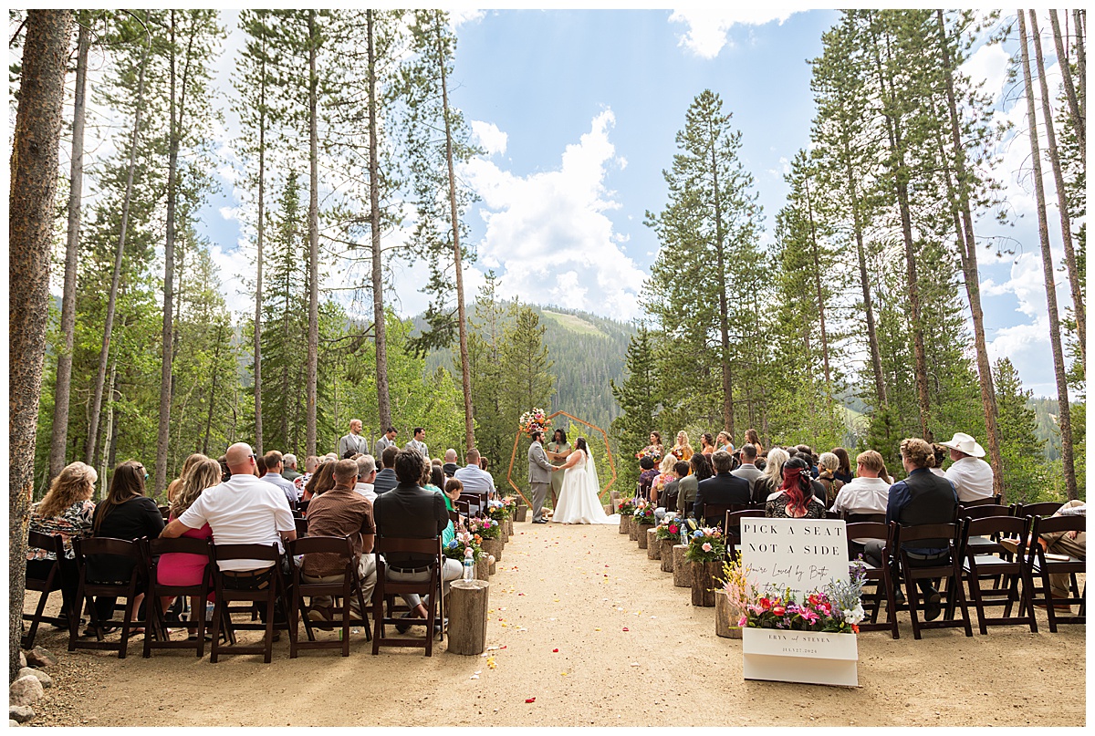 The bride and groom get married at Winter Park Mountain Lodge. They are holding hands. A heptagonal arch, pine trees, and a mountain are behind them.