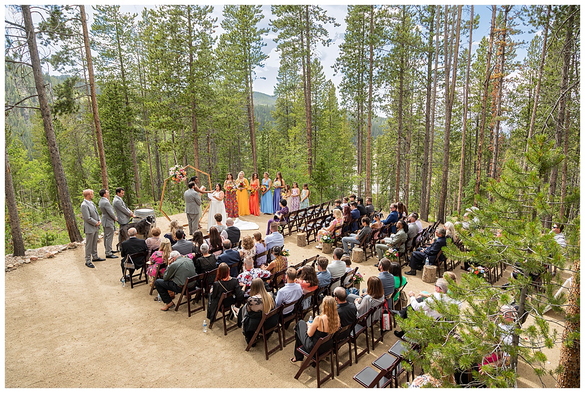 The bride and groom get married at Winter Park Mountain Lodge. The bride is reading her vows. A heptagonal arch, pine trees, and a mountain are behind them.