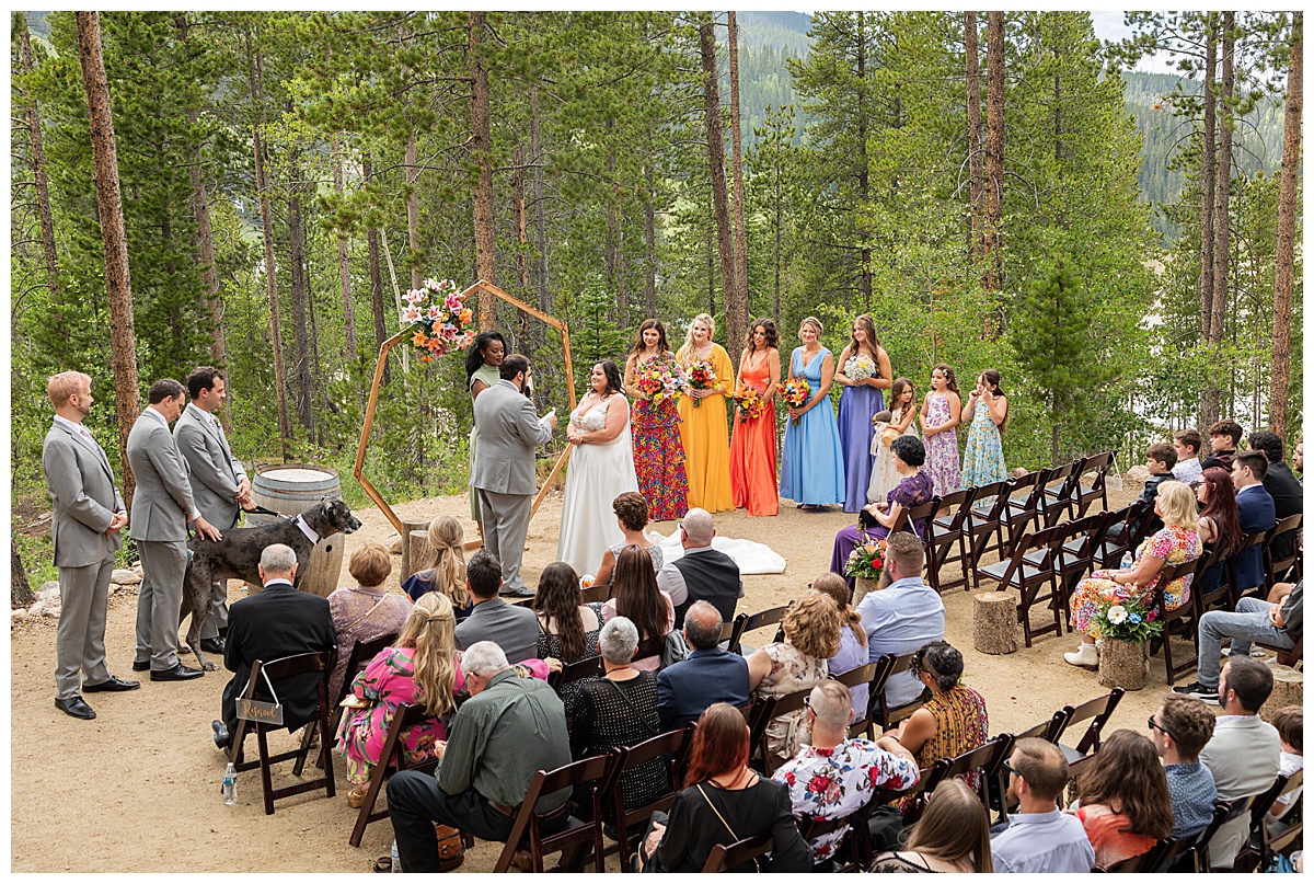 The bride and groom get married at Winter Park Mountain Lodge. The groom is reading his vows. A heptagonal arch, pine trees, and a mountain are behind them.