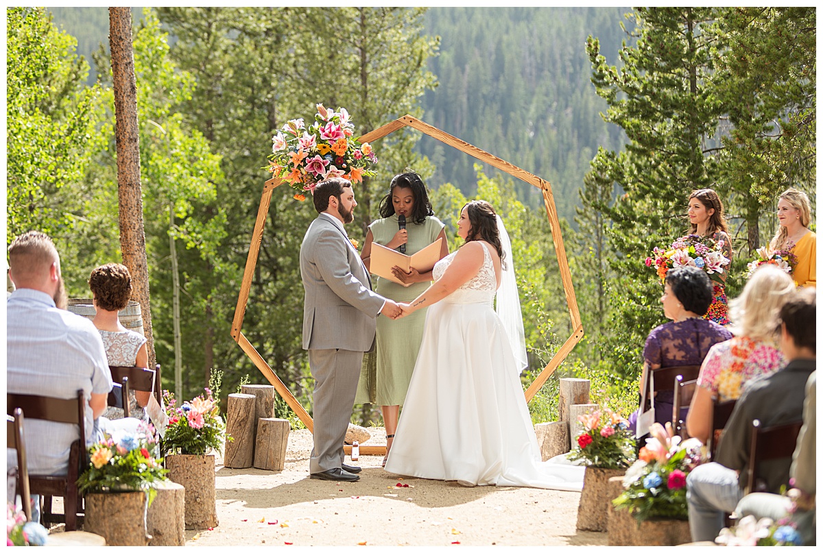 The bride and groom get married at Winter Park Mountain Lodge. They are holding hands. A heptagonal arch, pine trees, and a mountain are behind them.