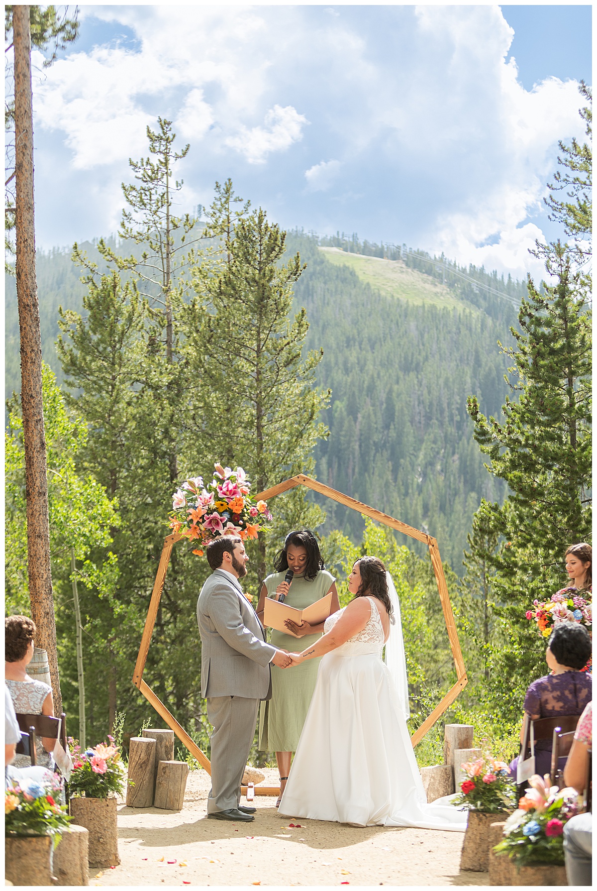 The bride and groom get married at Winter Park Mountain Lodge. They are holding hands. A heptagonal arch, pine trees, and a mountain are behind them.