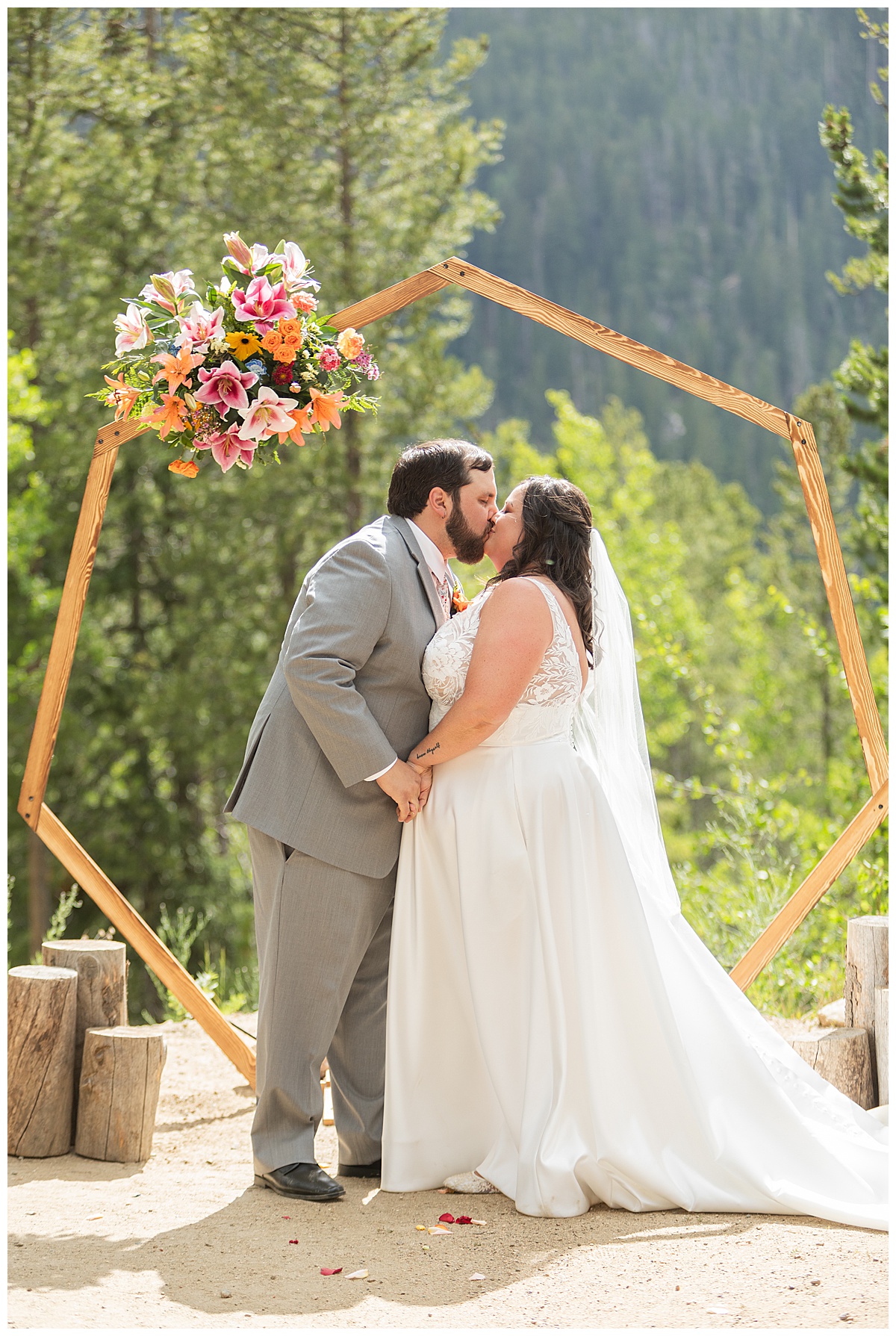 The bride and groom get married at Winter Park Mountain Lodge. They are having their first kiss. A heptagonal arch, pine trees, and a mountain are behind them.