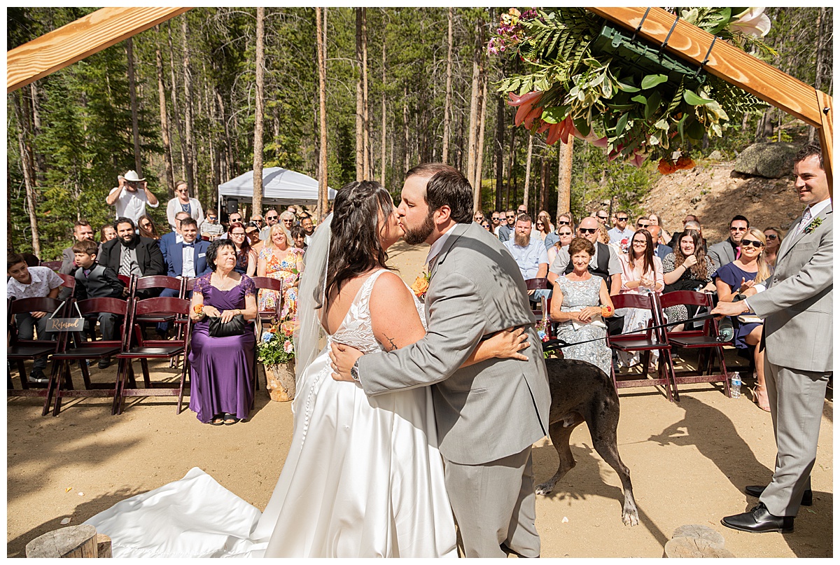 The bride and groom get married at Winter Park Mountain Lodge. They are having their first kiss. A heptagonal arch, pine trees, and a mountain are behind them.