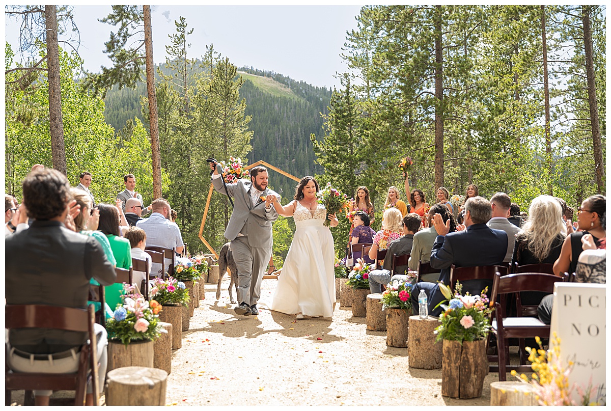 The bride and groom get married at Winter Park Mountain Lodge. They are walking down the aisle with Earl the great dane. A heptagonal arch, pine trees, and a mountain are behind them.