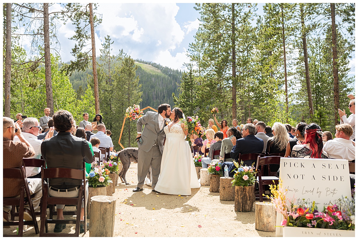 The bride and groom get married at Winter Park Mountain Lodge. They are walking down the aisle with Earl the great dane. A heptagonal arch, pine trees, and a mountain are behind them.