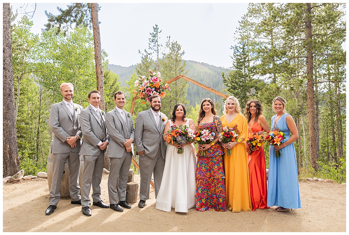 The bride, groom, and bridal party are posing for photos. The men are wearing gray suits and the women are in different colored dresses. Pine trees and a mountain are behind them.