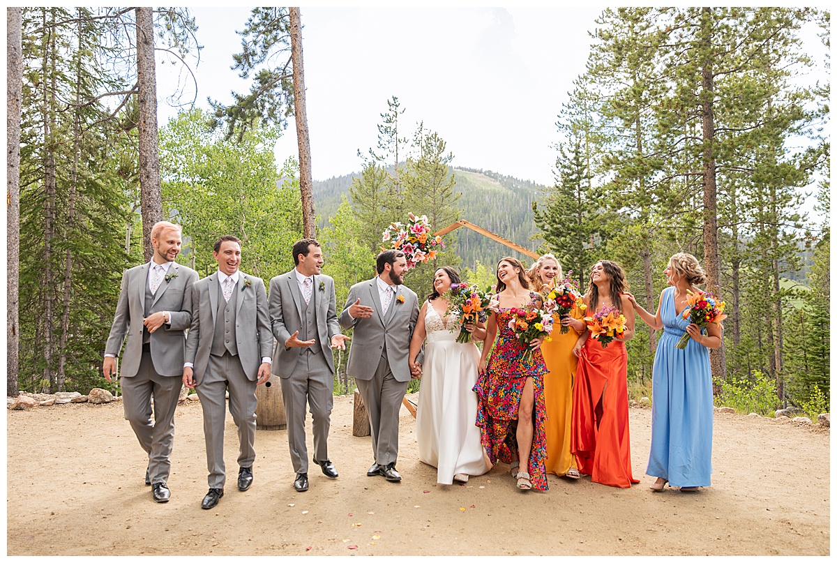 The bride, groom, and bridal party are posing for photos. The men are wearing gray suits and the women are in different colored dresses. Pine trees and a mountain are behind them.